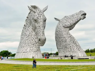 The Kelpies metal horse-head sculptures rising above a grassy field, with visitors near the base for scale.