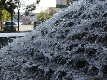 Cascading water over dark tiers with a street and parked cars blurred in the background near the National Katyń Memorial.