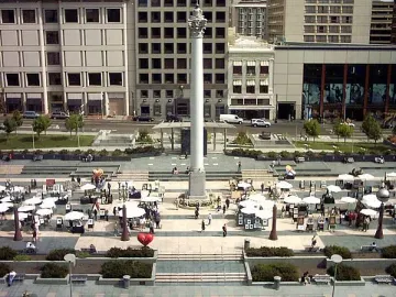 The Dewey Monument rises in the center of a busy public plaza with people gathered around its base and buildings in the background.