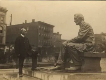 A man in a suit stands beside the Seated Lincoln statue with one foot on a low platform, looking toward the sculpture.
