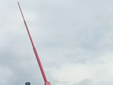 Prague Metronome mechanism with a red arm and counterweight against a cloudy sky.