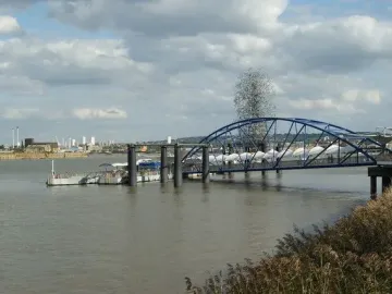 Quantum Cloud sculpture rises behind a blue bridge over the river near a pier.