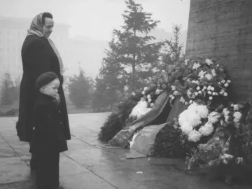 Floral wreaths and ribbons placed beside the Stalin monument, with a woman and child standing nearby.