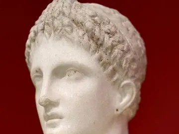 Marble bust of Artemis Diadoumena in profile against a red background, with short textured hair and draped shoulders.