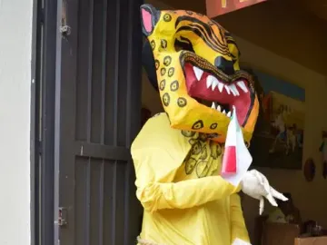 Mannequin wearing a yellow and black spotted jaguar mask, yellow jumpsuit, and white gloves, holding a small red, white, and green banner.