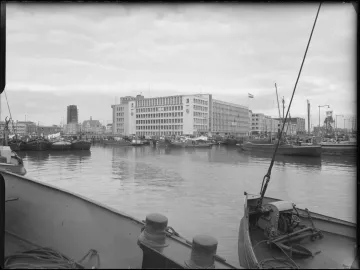 Boats moored in a harbor with multi-story buildings along the far bank under a cloudy sky.