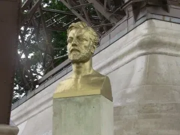 Golden bust of Gustave Eiffel on a stone pedestal with the Eiffel Tower framework in the background.