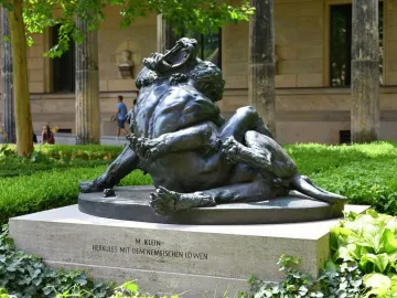 Dark bronze Hercules and the Nemean Lion statue on a stone pedestal beside a grand stone building.