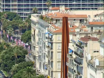 Neuf Lignes Obliques sculpture standing between apartment buildings on a busy city street lined with parked cars and palm trees.