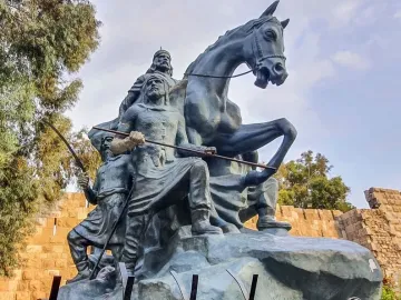 The Statue of Saladin, a bronze equestrian monument with two armed figures beside it, set against stone walls and trees.
