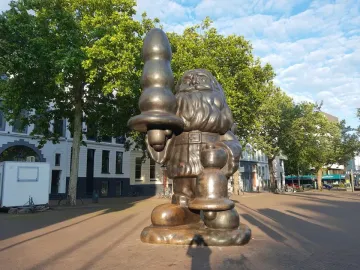 Bronze Santa Claus statue in a plaza, holding an abstract object in one hand and a bell-shaped object in the other.