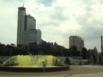 Silesian Insurgents\' Monument partly visible behind a fountain with yellow water jets and trees.