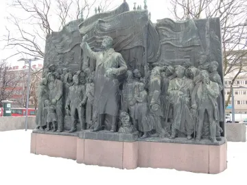 The Branting Monument in a snowy square, showing a bronze figure addressing a crowd with raised arm and draped flag-like forms behind.