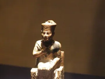 Khufu statuette of a seated figure wearing a tall crown on a clear base.