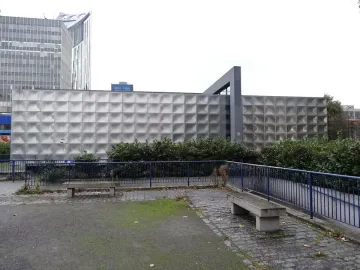 The Michael Faraday Memorial seen behind green bushes and a blue fence, with tall buildings in the background.