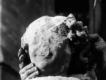 Damaged stone head of the Smiling Angel with a faint smile, held by two hands above a stone pedestal.