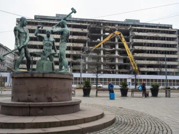 The Three Smiths bronze sculpture stands in front of a building demolition site with a yellow excavator in the background.