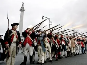 The Trenton Battle Monument rises above colonial reenactors firing muskets in the foreground.