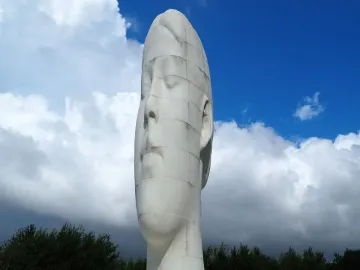 The Dream sculpture by Jaume Plensa, a tall white head and neck with closed eyes, against a blue sky with clouds.