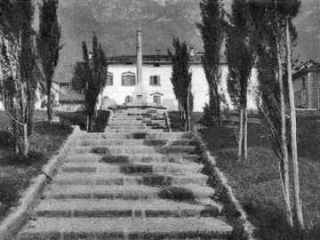 A long stone staircase leads up to the Lighthouse of Lierna, framed by slender trees with mountains in the background.