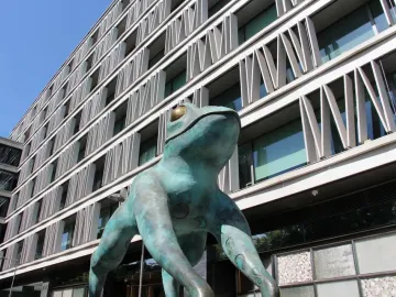 Lucky Frog statue with teal patina and golden eyes in front of a modern building.