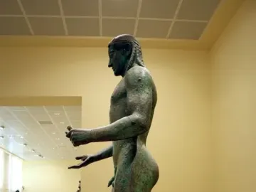 The Piraeus Apollo, a bronze nude male statue with braided hair, shown in profile on a stone base.