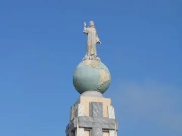 Divino Salvador del Mundo monument showing a robed figure raising one arm atop a globe and stone pedestal against a blue sky.