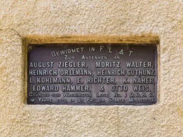 A bronze plaque with raised German text set into a light beige wall at the Hermann Heights Monument.