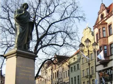 Nicolaus Copernicus Monument on a tall stone pedestal in a historic town square, with a fountain, benches, and old buildings nearby.