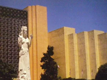 The Pacifica statue stands in a plaza, with raised arms, an elaborate headpiece, and people gathered at its base.