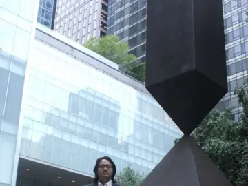 Broken Obelisk sculpture standing in front of modern glass buildings and greenery.