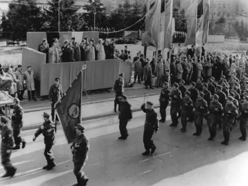 A statue of Joseph Stalin stands near a ceremonial parade with uniformed marchers, flags, and spectators on a raised platform.