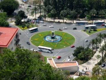 Three Graces Fountain in a traffic roundabout with water flowing into a blue basin and a star-shaped flowerbed around it.