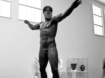 The Artemision Bronze stands on a plinth in a museum hall with arms extended.