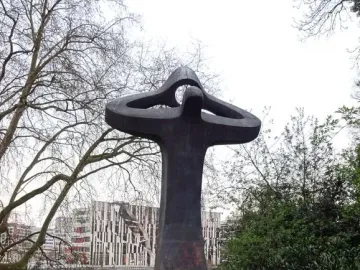 Der Mahner, a tall abstract sculpture with raised arms, stands on a lawn beside water and modern buildings.