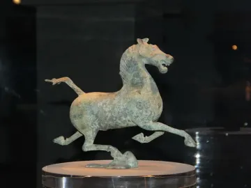 Weathered bronze Flying Horse of Gansu sculpture in a galloping pose on a pedestal inside a display case.