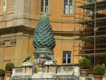 La Pigna bronze pine cone sculpture with peacocks on an ornate stone balustrade, in front of a building partly covered by scaffolding.