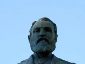 Weathered bronze bust of Maitland Brown in a suit and tie on a stone pedestal against a blue sky.