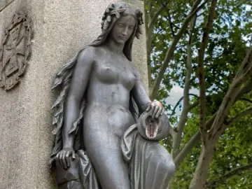 Mutter Kinzig statue with a leafy wreath, leaning against a stone monument and holding an overturned urn.