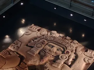 Tlaltecuhtli Monolith displayed flat in a museum, showing a carved face, headdress, and weathered stone surface.