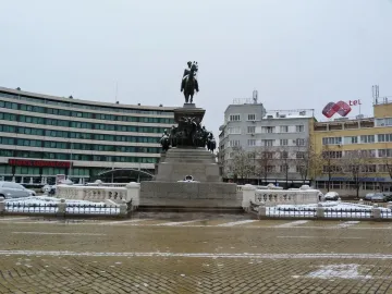The Tsar Liberator Monument stands on an ornate pedestal in a wide square, with a classical building and domed structure behind it.