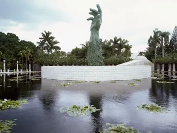 Holocaust Memorial sculpture of a towering bronze hand formed by human figures, rising beside a white curved wall and reflecting pool with palm trees nearby.