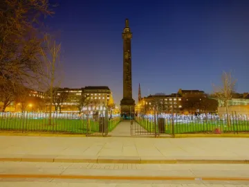The Melville Monument rises above glowing lights in St Andrews Square at twilight, with illuminated buildings in the background.