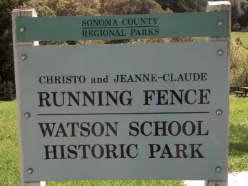 Light-green and white historical site sign for Running Fence on a grassy lawn with trees behind it.