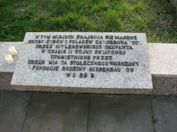 Granite memorial plaque for the Monument of Common Martyrdom with raised Polish lettering and two white candles beside it.