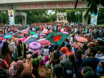 Anti Terrorism Raju Memorial Sculpture beside a large crowd with umbrellas, Bangladeshi flags, and a concrete archway in the background.