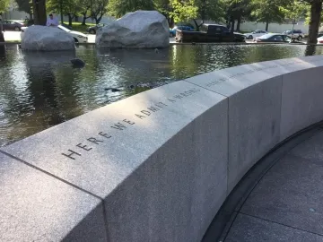 Curved stone wall of the Japanese American Memorial inscribed with “HERE WE ADMIT A WRONG,” beside a reflecting pool and boulders.