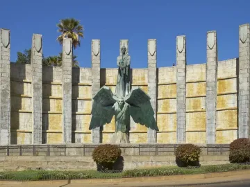 Monumento a Franco with a bronze robed figure holding a sword atop a winged figure in front of a curved stone wall and pillars.