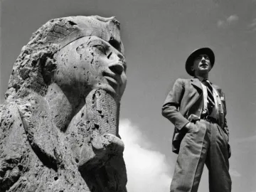 The Sphinx of Memphis with a man in a suit and fedora standing beside it under a cloudy sky.