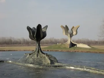 A dark Stone Flower sculpture in the foreground with the larger Kameni cvijet monument across the water on a grassy mound.